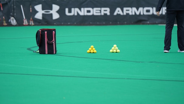 A sports field features a black and pink sports bag, two pyramids of yellow balls, and a portion of a person in dark athletic gear. The background displays a banner with the Under Armour logo.