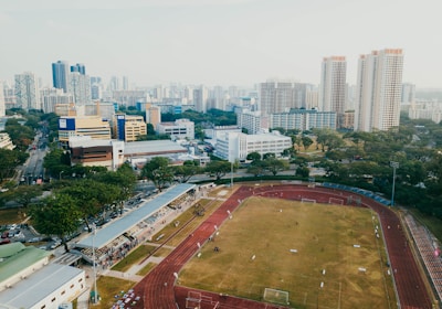 An aerial view of a sports complex with a running track surrounding a grassy field, set against the background of a cityscape. The city's skyline features a mix of tall residential buildings and shorter commercial structures. There are trees and green areas surrounding the sports facility.