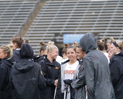 A group of people, likely athletes, are gathered together on a sports field. They are wearing black jackets with some individuals dressed in white sports uniforms underneath. A few people have their hair tied up in buns, and most are engaged in conversation. The background shows tiered stadium seats, suggesting the setting is an outdoor sports venue.
