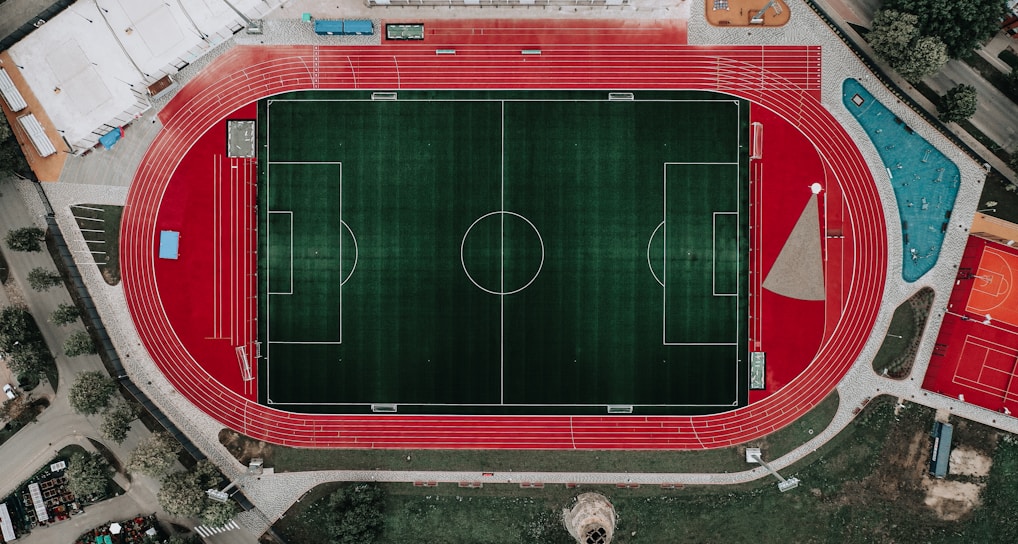 An aerial view of a large sports complex featuring a central green football field surrounded by a red running track. Adjacent to the track, there is a parking area, buildings, and pathways. Nearby, there are additional sports courts in different colors, including a blue swimming pool and orange basketball courts.