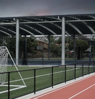 An outdoor sports facility featuring a running track and a soccer goal. The track is red, surrounded by a black metal fence, and the field is covered with artificial green turf. In the background, a basketball court is visible under a covered roof with metal supports. The sky is overcast, adding a moody atmosphere.
